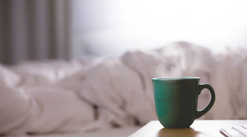 A green coffee cup on a bedside table, bright bedsheets bundled on the bed in the background.