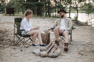 A couple in camping chairs drinking coffee by a riverbank.