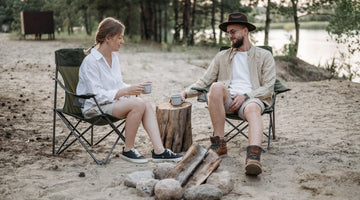 A couple in camping chairs drinking coffee by a riverbank.