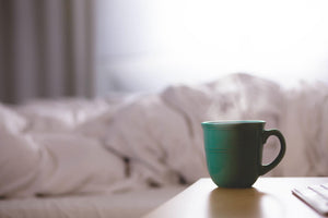 A green coffee cup on a bedside table, bright bedsheets bundled on the bed in the background.