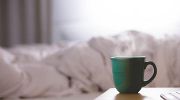 A green coffee cup on a bedside table, bright bedsheets bundled on the bed in the background.