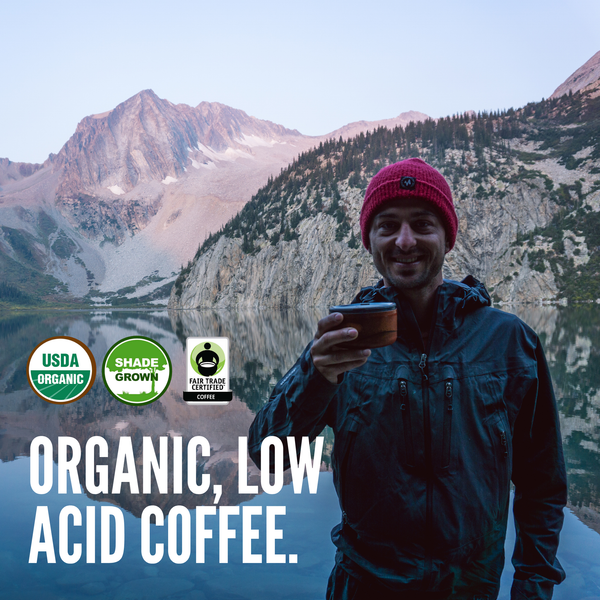 A smiling man in a red beanie and dark jacket stands in front of a mountain lake holding a coffee cup, with USDA Organic, Shade Grown, and Fair Trade Certified logos and the text 'ORGANIC, LOW ACID COFFEE.' overlaid on the image.