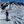 Load image into Gallery viewer, A person cross-country skiing with poles on a snowy mountain trail, accompanied by a light-colored dog, with snow-covered mountains and a blue sky in the background.
