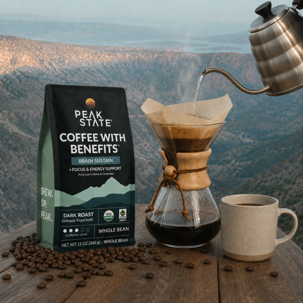 A bag of Peak State Coffee with Benefits is shown on a wooden table with coffee beans scattered around it, alongside a glass pour-over coffee maker, a ceramic mug of coffee, and a metal kettle pouring water, all set against a blurred mountain landscape background.